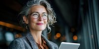 Confident mature businesswoman with silver curly hair and glasses holding digital tablet, smiling against blurred office background with bokeh lights.