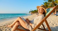 Young woman relaxing in beach chair reading book on tropical sandy shore with palm trees and turquoise ocean water during sunny vacation day.