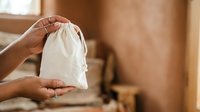 Close-up of hands holding a small white fabric pouch or drawstring bag against a blurred warm interior background, suitable for product packaging, storage solutions, and retail presentations.