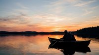 Silhouetted male figure sitting alone in small rowboat on calm lake at sunset, golden orange sky reflecting on water surface, forested shoreline in background, peaceful evening scene.