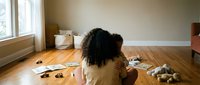 Young African American woman sitting on hardwood floor playing with adorable puppies in bright sunlit room. Pet adoption, animal care and companionship concept.