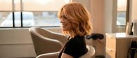 Professional blonde woman laughing joyfully in modern office chair, expressing happiness and positive workplace emotions in bright corporate environment.