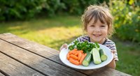 Happy young girl with blonde hair sitting at wooden table outdoors presenting plate of fresh vegetables including carrots and cucumbers in sunny garden setting.