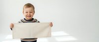Happy toddler boy holding blank white paper sign with cheerful smile, wearing striped shirt against neutral background for advertising mockup.