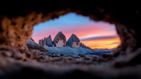 Dramatic sunset view of snow-covered Tre Cime di Lavaredo peaks through natural rock window frame in Dolomites. Vibrant orange and pink sky creates magical mountain landscape.