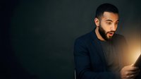 Young Middle Eastern man with dark beard wearing navy blazer and black shirt, illuminated by warm studio lighting against dark background, suitable for business, corporate, and professional portraits.