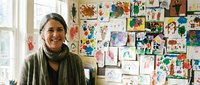 Smiling middle-aged Caucasian woman art teacher standing in front of colorful children drawings displayed on wall in bright classroom.