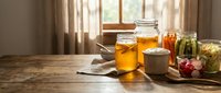 Homemade preserves and pickled vegetables in glass jars on rustic wooden table with natural sunlight streaming through kitchen window creating warm atmosphere.