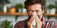 Middle-aged Caucasian man with beard sneezing into tissue, wearing plaid shirt in home kitchen setting with blurred green plants in background.