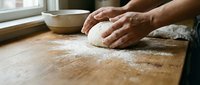Hands kneading fresh dough on wooden cutting board with flour in warm kitchen setting for homemade bread baking and cooking preparation.