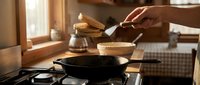 Person cooking pancakes in cast iron skillet on stovetop in warm rustic kitchen with natural lighting for breakfast preparation and home cooking concepts.