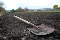 Old rustic shovel with wooden handle lying on freshly plowed dark soil in rural agricultural field, farming equipment against countryside landscape.