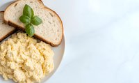 Creamy scrambled eggs served on white plate with fresh bread slices and basil garnish on marble background, bright morning breakfast setup with copy space.
