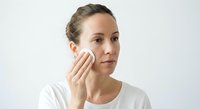 Young Caucasian woman applying skincare cream to face with cotton pad in white shirt against neutral background for beauty and wellness concepts.
