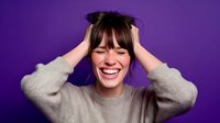 Young woman with hands in hair laughing joyfully against purple background, expressing happiness and carefree attitude in casual gray sweater.