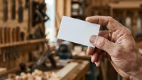 Close-up of male hand holding blank white business card at construction site with wooden materials and tools in blurred background.