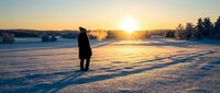 Solitary figure stands on frozen lake at sunrise, surrounded by snow-covered landscape and golden sunlight reflecting off ice surface, winter solitude and nature exploration.