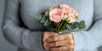 Elderly woman's hands holding delicate pink roses and eucalyptus bouquet against gray sweater background, representing care, aging and tenderness in panoramic crop.