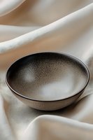 Minimalist ceramic bowl with speckled black glaze on cream fabric background, showcasing artisanal craftsmanship and modern design aesthetic for home decor.