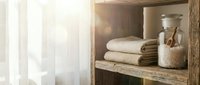 Rustic wooden shelf displaying neatly folded cream and olive towels with glass jar containing bath salts, bathroomorganization and home decor.