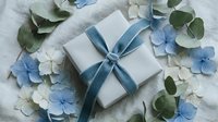 White gift box with blue satin ribbon surrounded by delicate blue and white hydrangea flowers and eucalyptus leaves on soft fabric background.