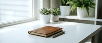 Minimalist workspace with leather notebook and pen on white desk near window with green plants creating productive home office environment.