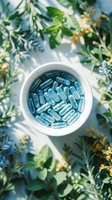 Blue capsules in white bowl surrounded by fresh green herbs and flowers for natural health and wellness supplement concept.