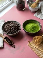 Coffee beans and matcha green tea powder in wooden bowls with traditional bamboo whisks on pink surface for healthy beverage preparation and cooking.