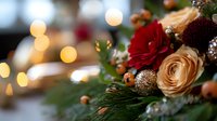 Romantic floral arrangement with red and peach roses, evergreen branches and gold ornaments against soft bokeh lights, creating festive holiday atmosphere.
