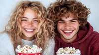 Young couple with curly hair laughing while eating popcorn together, casual lifestyle portrait showing joy and friendship on white background.
