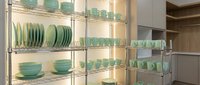 Mint green ceramic dishes, bowls, and mugs neatly arranged on illuminated metal shelves in a modern kitchen pantry storage area.