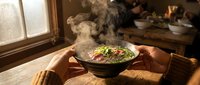 Hands holding steaming bowl of noodles with vegetables in warm kitchen setting, steam rising from hot Asian food dish, comfort meal preparation.