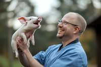Cheerful male veterinarian in blue scrubs holding playful pink piglet outdoors, expressing joy and professional care while interacting with young farm animal.