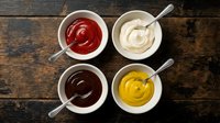 Four colorful condiment sauces in white bowls with spoons on rustic wooden table. Ketchup, mayo, chocolate and mustard dips for cooking and dining.