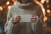 Woman in cozy knit sweater holding blank white card against warm bokeh lights background, creating atmospheric holiday mood for winter greetings or messages.