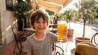 Happy young boy enjoying fresh orange juice at outdoor cafe terrace during sunny morning breakfast time with family vacation vibes.