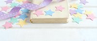 Colorful paper stars scattered around closed book on white wooden surface creating festive educational concept for learning celebration and achievement.