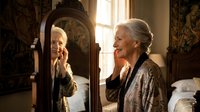 Elderly Caucasian woman with gray hair looking at herself in vintage mirror, touching face in contemplative moment. Warm natural lighting creates intimate atmosphere.