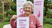 Senior Caucasian woman breast cancer survivor holding awareness sign with pink ribbon during National Cancer Survivor Month outdoor health campaign event.