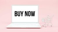 White laptop with BUY NOW text on screen next to a miniature shopping cart on a pink background, symbolizing online shopping and e-commerce.