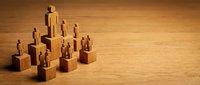 Wooden human figures standing on blocks of varying heights representing hierarchy, teamwork and organizational structure on natural wood surface.