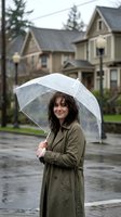 Young woman with dark hair and fair complexion holding transparent umbrella on rainy residential street. Wearing olive green trench coat. Suburban neighborhood with two-story houses and evergreen trees in background. Wet pavement and overcast weather conditions.
