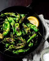 Grilled shishito peppers served on black plate with lemon wedges and sea salt. Charred green peppers with blistered skin create appetizing contrast in dark moody setting.