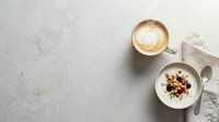 Overhead view of cappuccino with latte art and granola yogurt bowl on gray concrete surface, ideal for cafe, breakfast, and food styling projects.