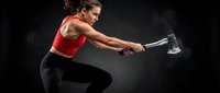 Athletic woman with curly hair swinging sledgehammer in dynamic fitness training pose against dark background for strength workout programs.