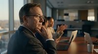 Mature male professional with glasses in thoughtful pose at laptop during office meeting, suitable for business, corporate training, leadership, and workplace communication concepts.