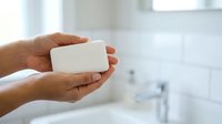 Hands holding white soap bar in bathroom with blurred sink and mirror background for hygiene and cleanliness concepts.
