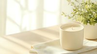 Minimalist white scented candle in ceramic holder on linen napkin with green plant and soft window light creating cozy atmosphere for spa and relaxation.