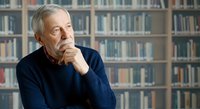 Senior man with gray hair and Caucasian ethnicity wearing dark blue sweater, hand on chin in thoughtful pose, sitting in library with bookshelves background.