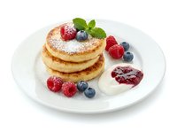 Stacked fluffy pancakes dusted with powdered sugar, topped with fresh raspberries and blueberries, served with whipped cream and berry jam on white plate.